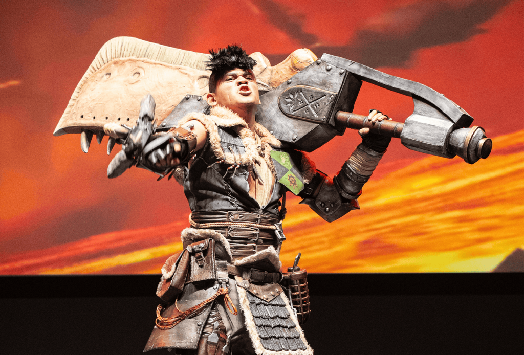 Cosplayer trabajando detalladamente en una armadura futurista dentro de un taller lleno de herramientas y bocetos, con el logo de CCXP México de fondo.