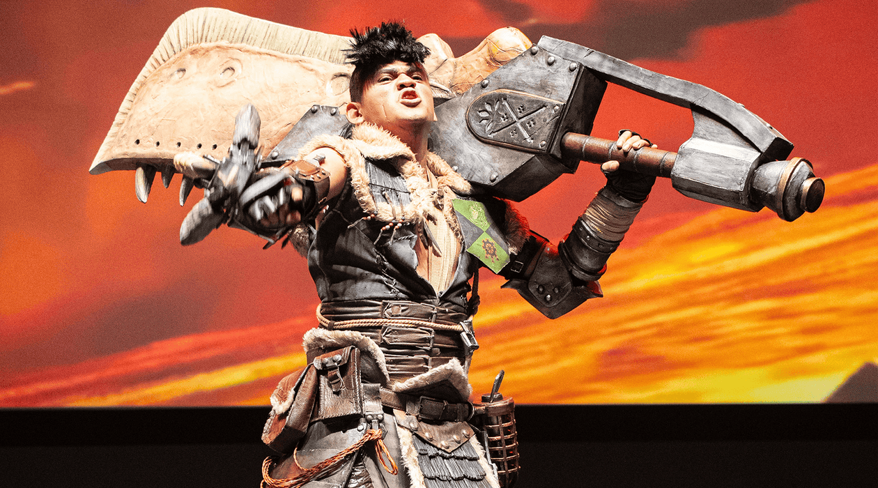 Cosplayer trabajando detalladamente en una armadura futurista dentro de un taller lleno de herramientas y bocetos, con el logo de CCXP México de fondo.