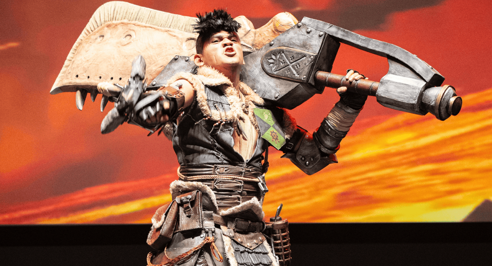 Cosplayer trabajando detalladamente en una armadura futurista dentro de un taller lleno de herramientas y bocetos, con el logo de CCXP México de fondo.